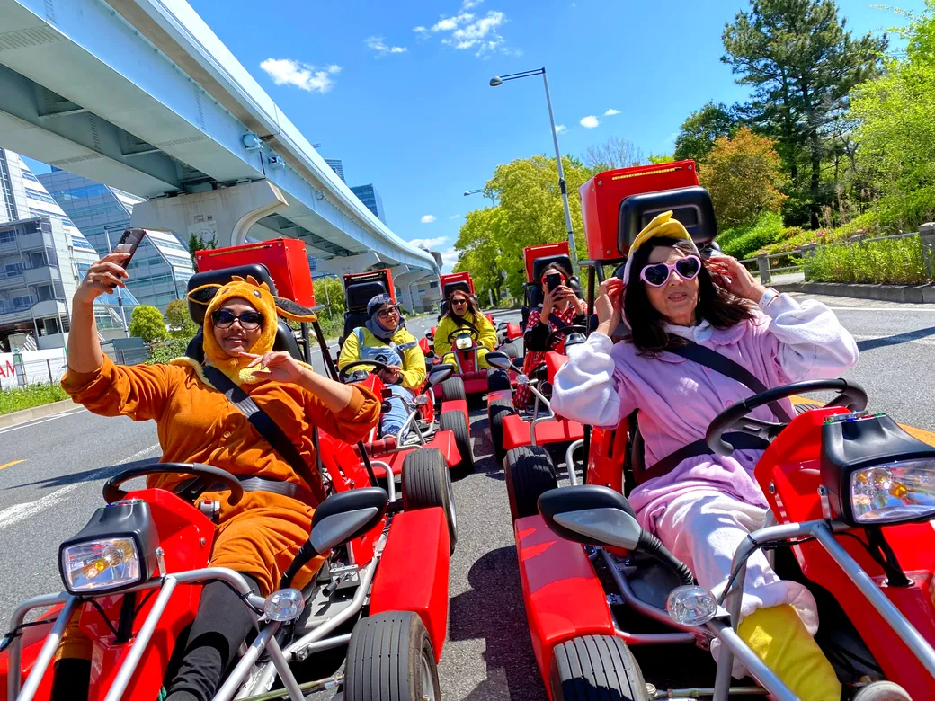 Group of people in bright red go-karts wearing colorful animal costumes and sunglasses, taking a selfie on a sunny city street under an overpass.