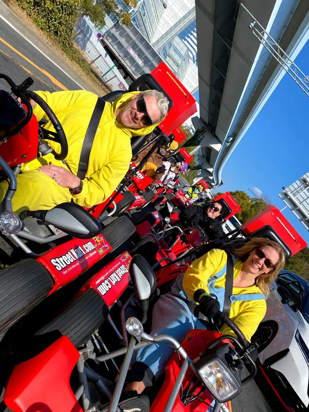 Group of people riding bright red go-karts in a line on a city street, wearing yellow jackets and sunglasses.