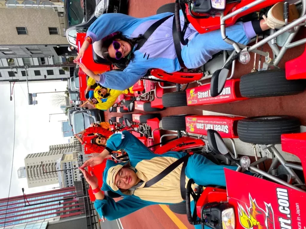 Several people in blue outfits pose in red go-karts lined up on a city street, waving and smiling at the camera.