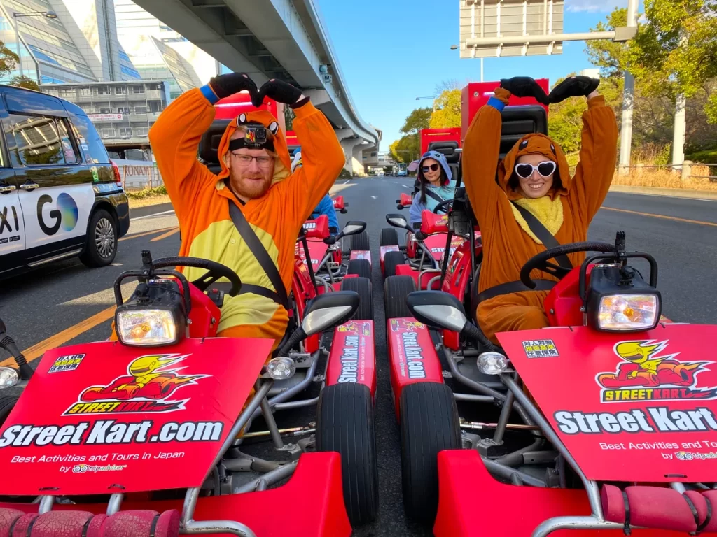 IMG_3605_converted - StreetKart Three people in bright orange animal onesies drive red Street Kart go-karts on a city street, posing with hands raised in the air.